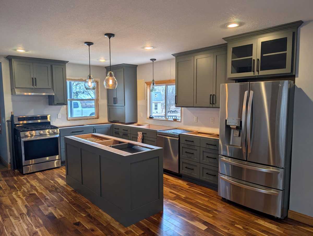 Deep green shaker kitchen with an island, crown molding, glass-front cabinets over the refrigerator and black pulls