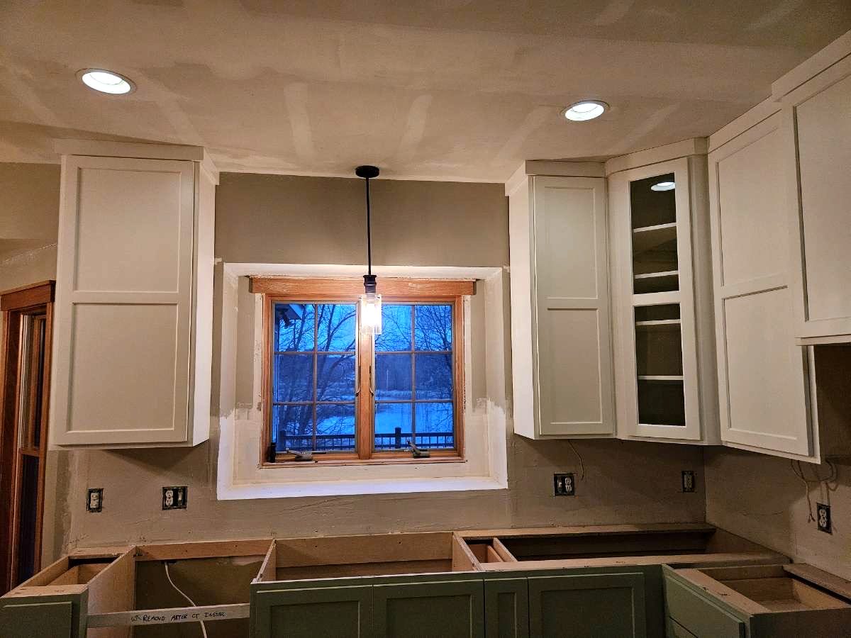 White upper cabinets flanking a kitchen window with a pendant light, above sage green base cabinets